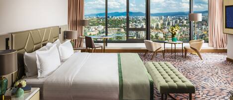 A hotel room with a large bed, a desk, a chair, a small table, and a view of a cityscape through large windows.