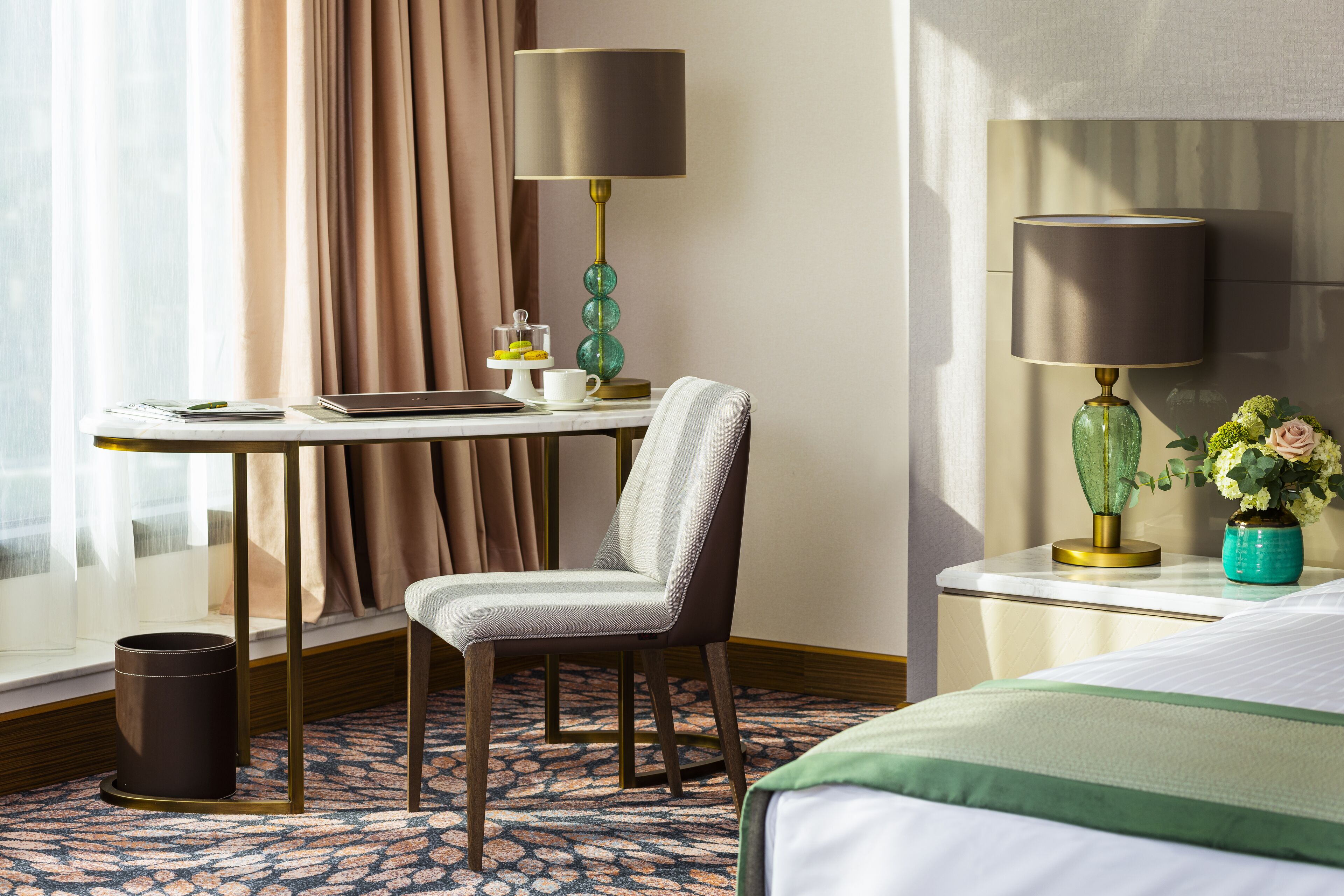 A hotel room with a bed, a desk with a laptop, a chair, a lamp, a vase with flowers, and a window with curtains.
