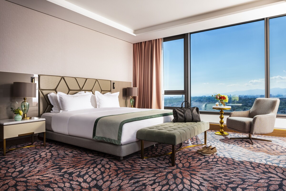 Une chambre d’hôtel moderne dotée d’un grand lit, d’un coin salon avec un fauteuil et une petite table, et offrant une vue sur le paysage urbain grâce à de grandes fenêtres.