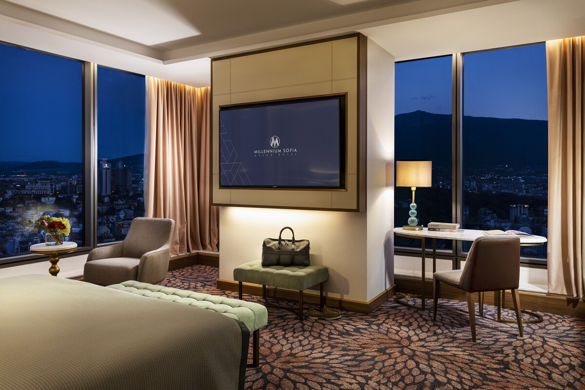 Une chambre d’hôtel avec un grand lit, un bureau avec une lampe, une chaise, une petite table avec un vase et un téléviseur affichant le nom de l’hôtel.
