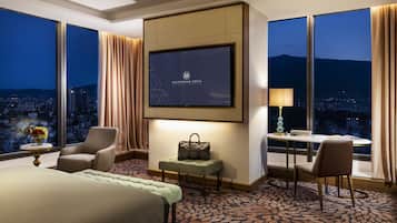 Une chambre d’hôtel avec un grand lit, un bureau avec une lampe, une chaise, une petite table avec un vase et un téléviseur affichant le nom de l’hôtel.