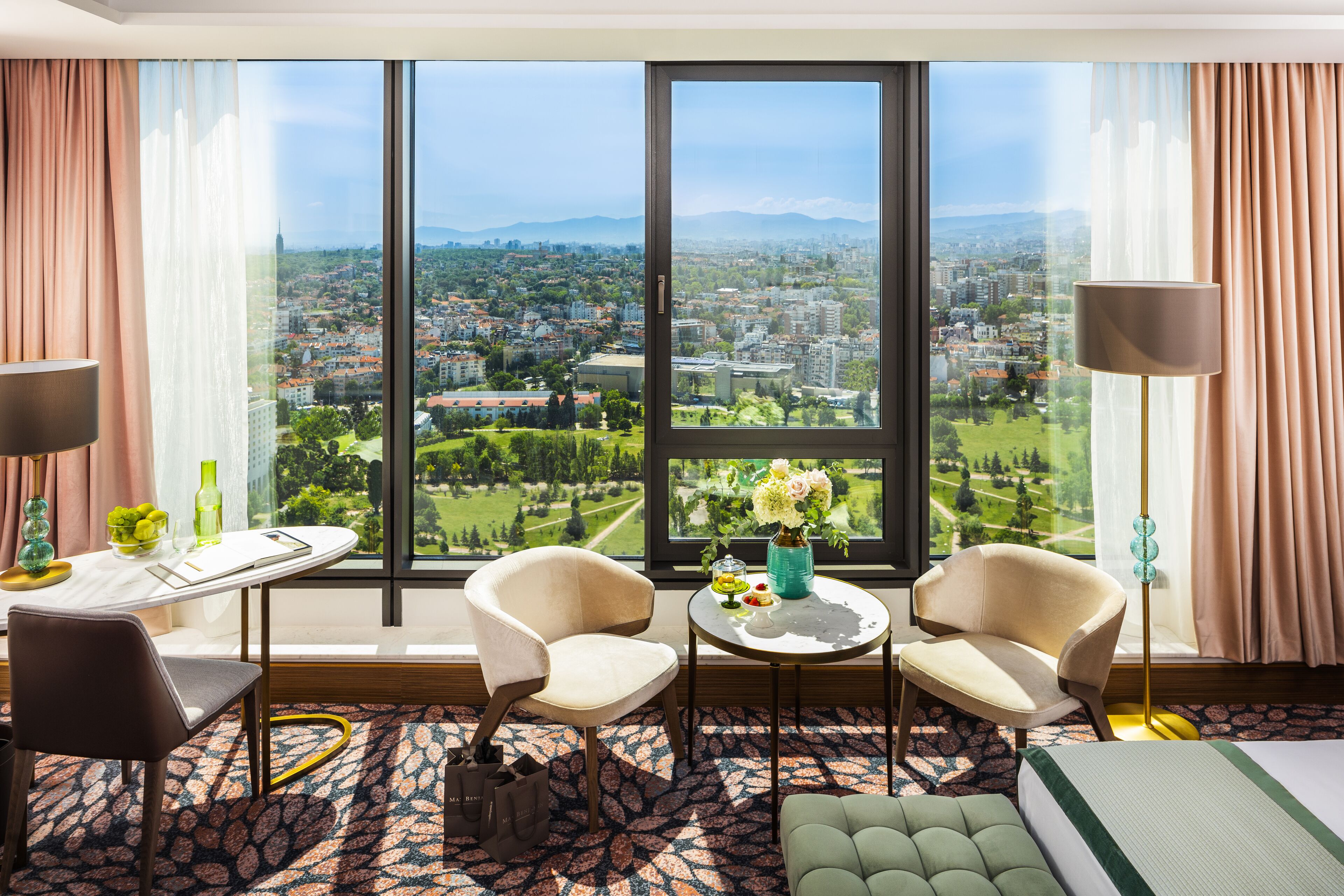 A hotel room with a large window offering a city view, two armchairs, a small table with a vase, and a bed.