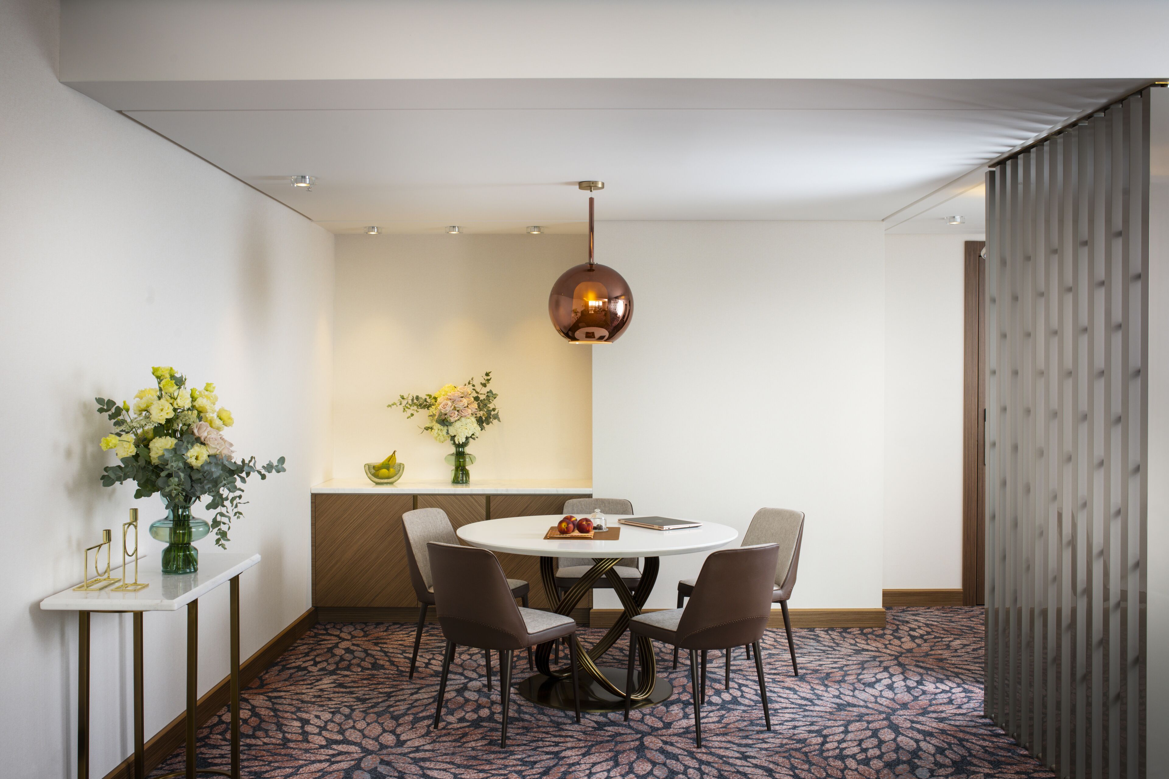 Un comedor con una mesa redonda, cuatro sillas, un florero con flores y una lámpara colgante.