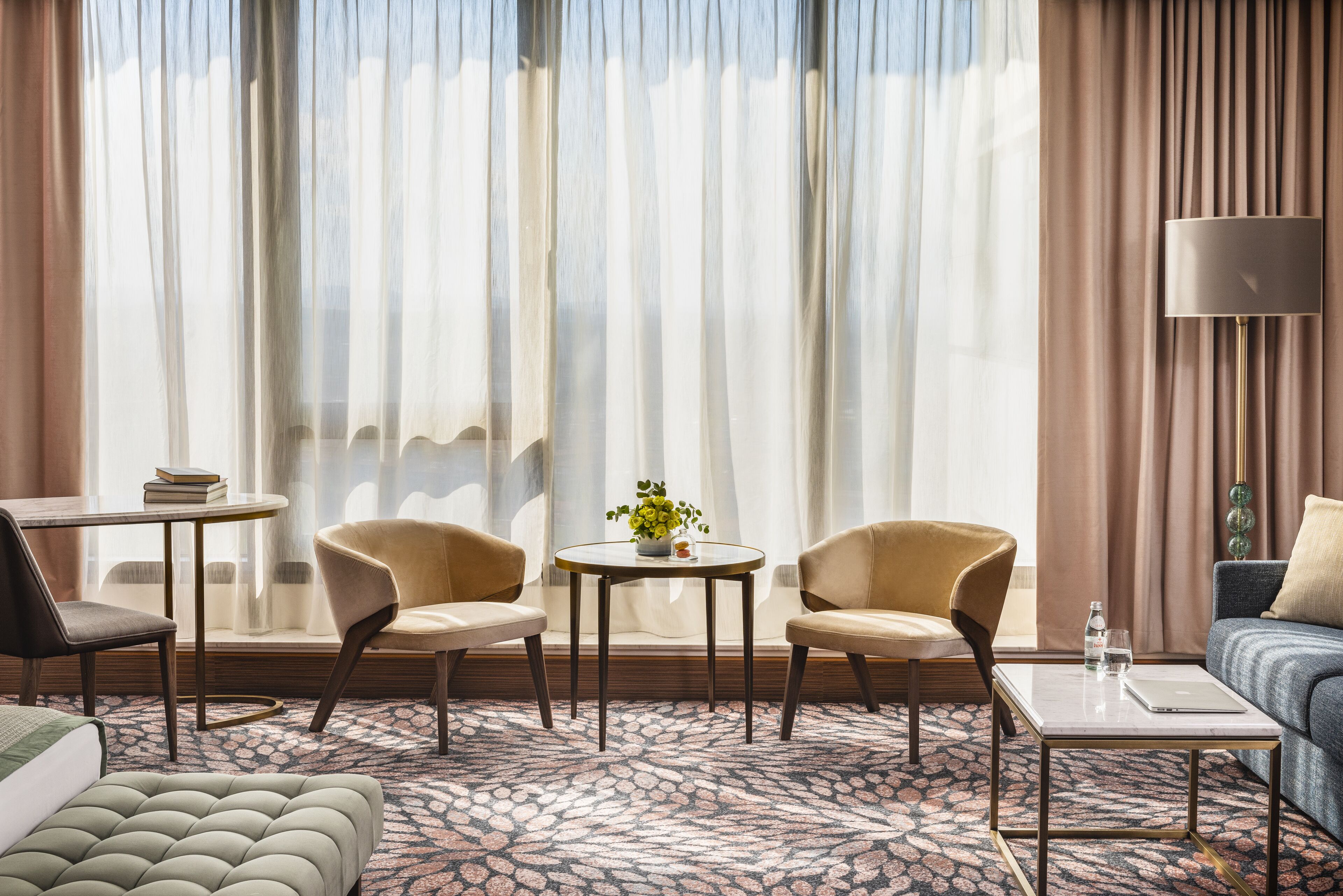 Habitación de hotel con un ventanal grande, dos sillones, una mesa redonda con una planta, un sofá, una mesita con un ordenador portátil y una lámpara.