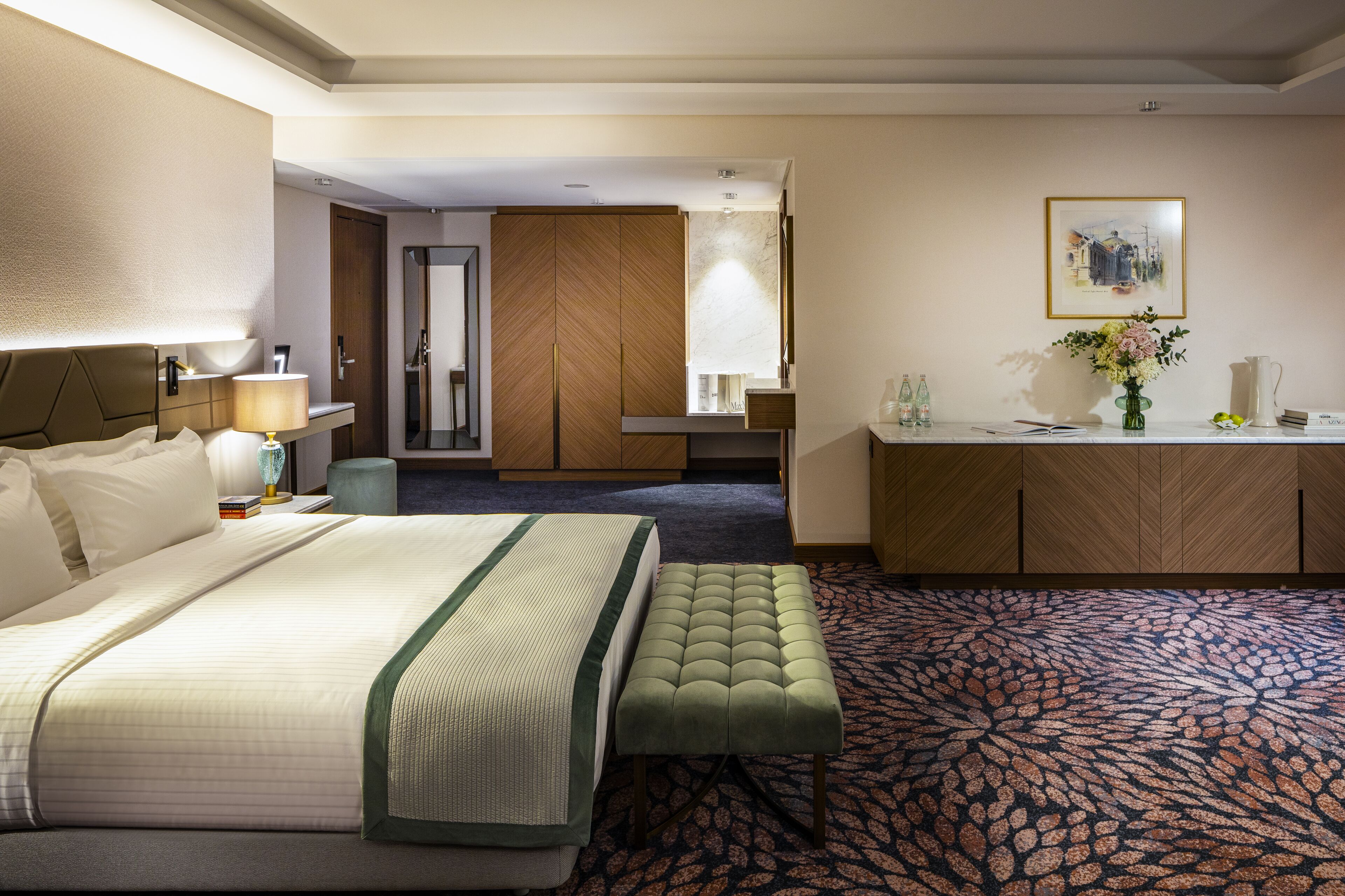 A hotel room with a large bed, a green bench, a desk, a mirror, and a framed picture on the wall.