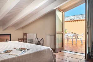 Un dormitorio con cama, una silla, un radiador y una puerta abierta que da a un patio exterior con mesa y sillas.