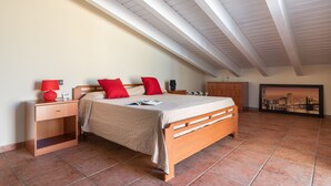 A bedroom with a sloped ceiling, a wooden bed, a nightstand with a lamp, a dresser, and a framed cityscape picture on the wall.