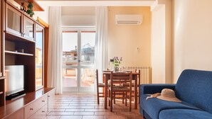 A living room with a blue sofa, wooden dining table, and chairs. There is a TV on a wooden cabinet, and an air conditioning unit on the wall.