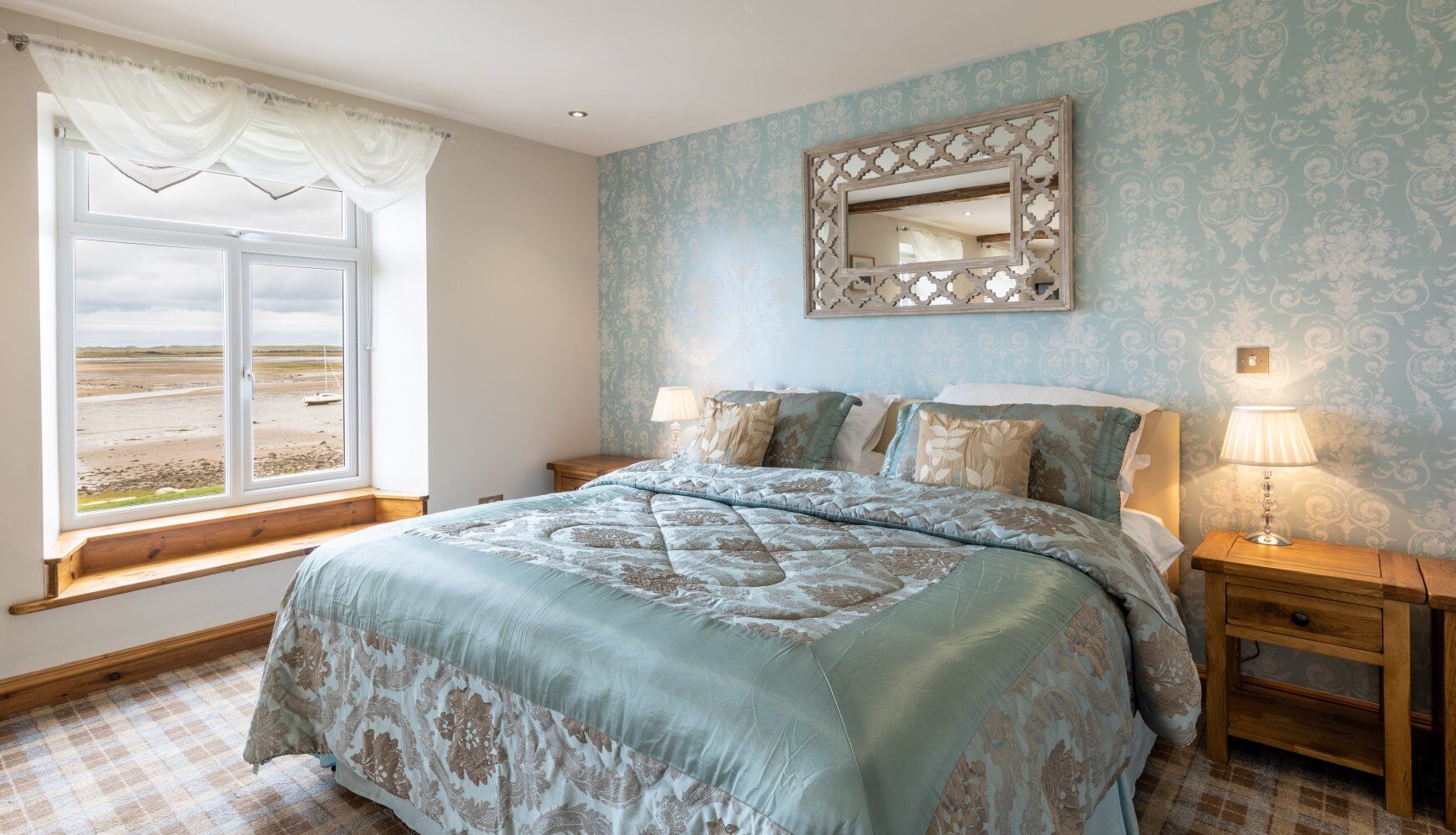 A bedroom with a large bed, a window with a view of the beach, a mirror on the wall, and a bedside table with a lamp.