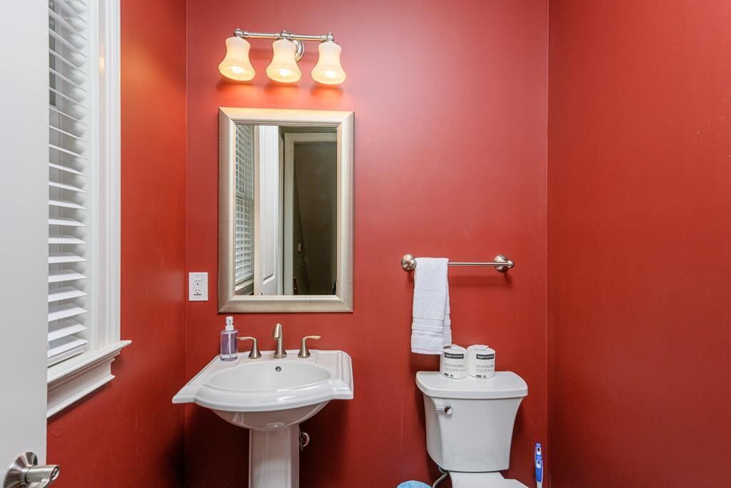 Une salle de bain avec un mur rouge, un lavabo blanc et des toilettes. Il y a un miroir au-dessus du lavabo et un porte-serviettes sur le mur.