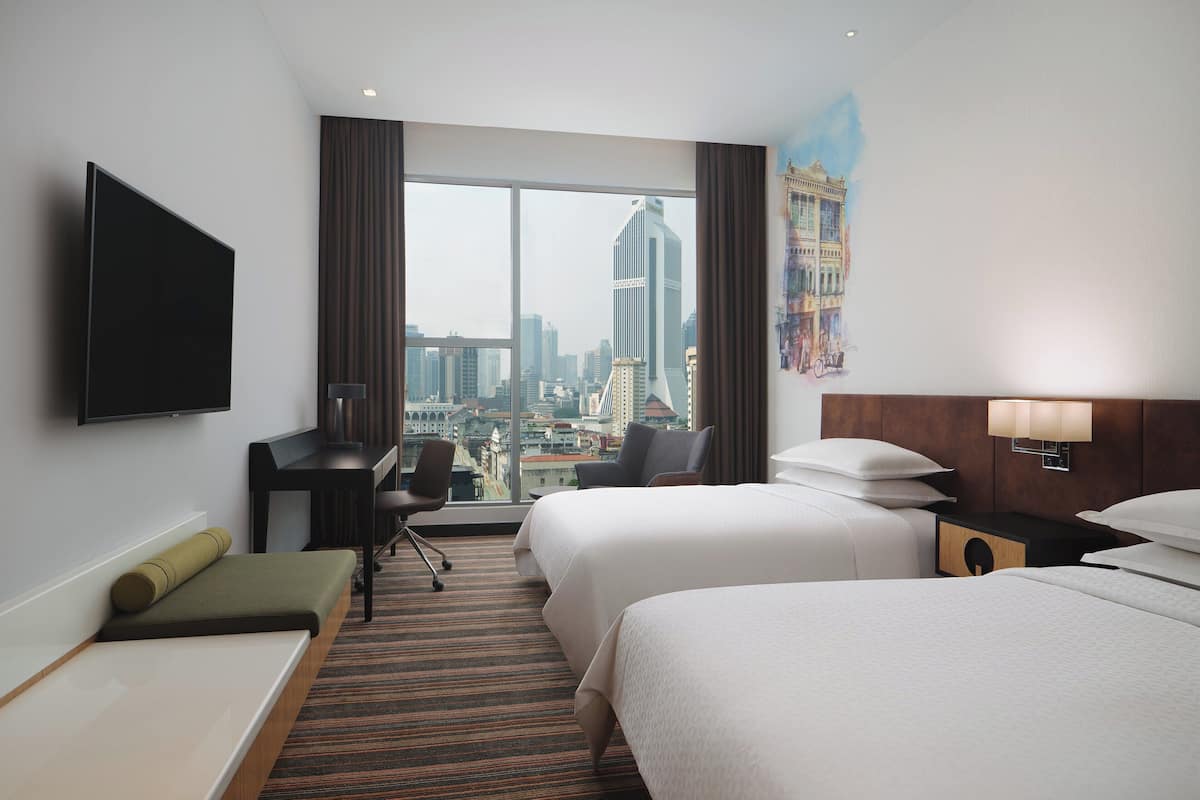 Une chambre d’hôtel avec deux lits, un bureau, une chaise, une télévision et une grande fenêtre donnant sur la ville.