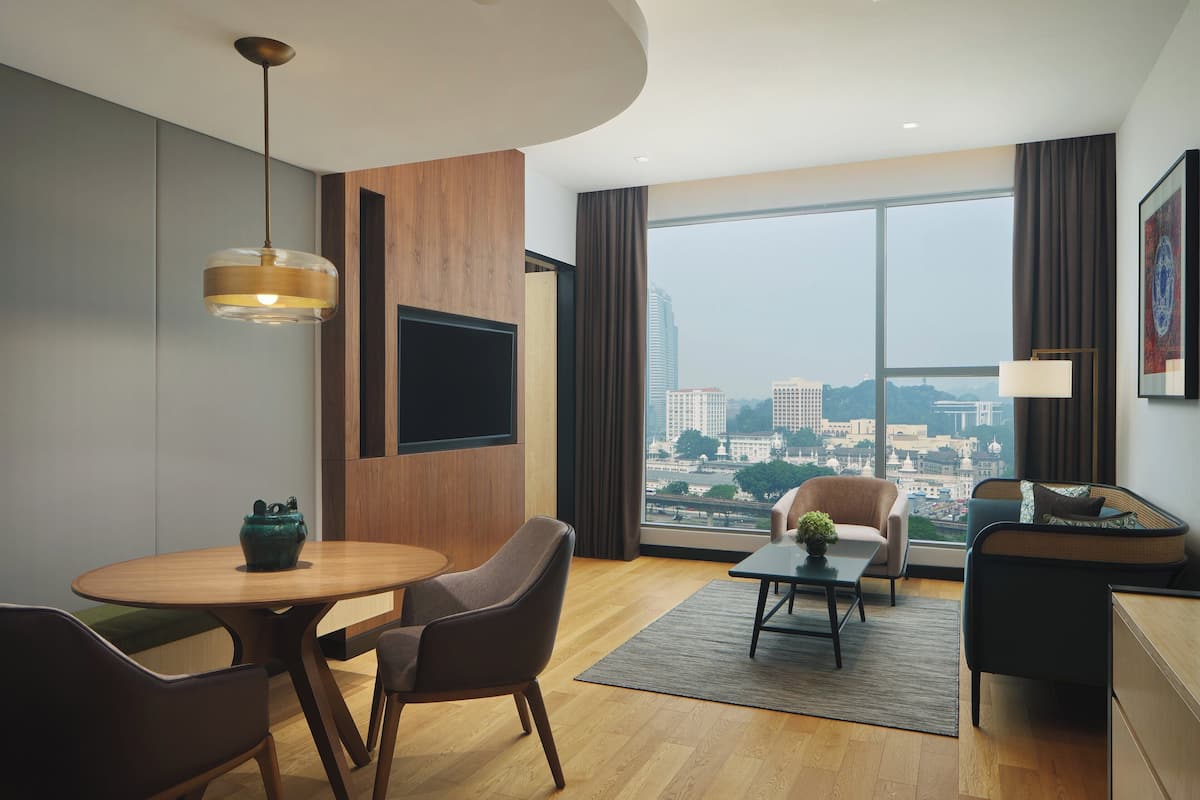 Une chambre d’hôtel moderne avec une grande fenêtre, une table à manger ronde, deux chaises, un canapé et un téléviseur fixé au mur.