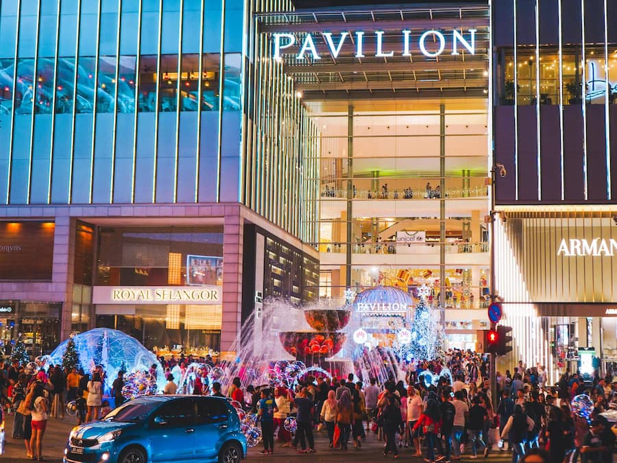 Una calle urbana concurrida, con gente, un coche azul y tiendas que incluyen 'PAVILION' y 'ARMANI'.