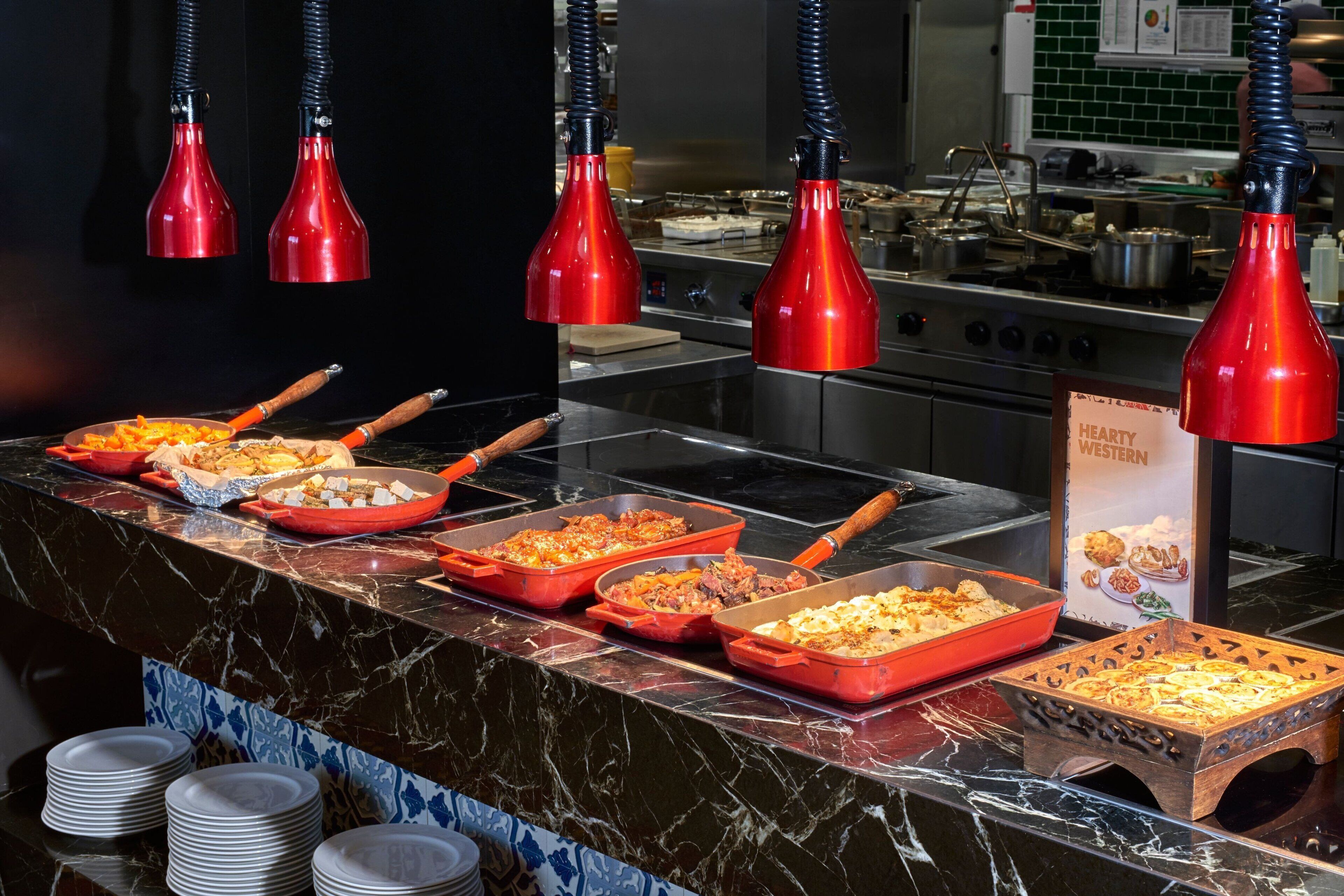 Ein reichhaltiges Buffet mit verschiedenen Speisen, roten Pendelleuchten und einem Schild, das 'HEARTY WESTERN' Küche anzeigt.
