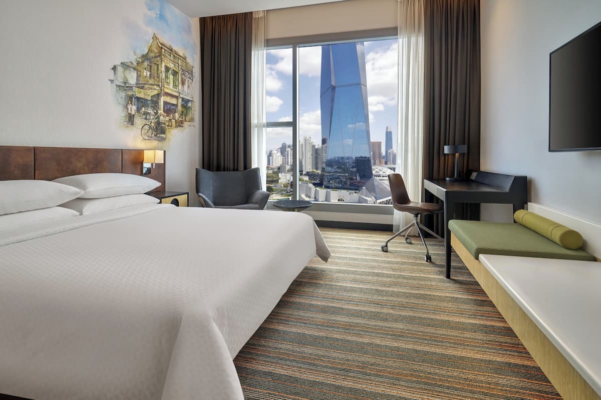 Habitación de hotel con una cama grande, un escritorio, una silla y vistas a un paisaje urbano a través de la ventana.
