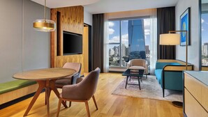 Habitación de hotel moderna con mesa de comedor de madera, asientos cómodos, televisor de pantalla plana, sofá, escritorio y una ventana grande con vistas a la ciudad.