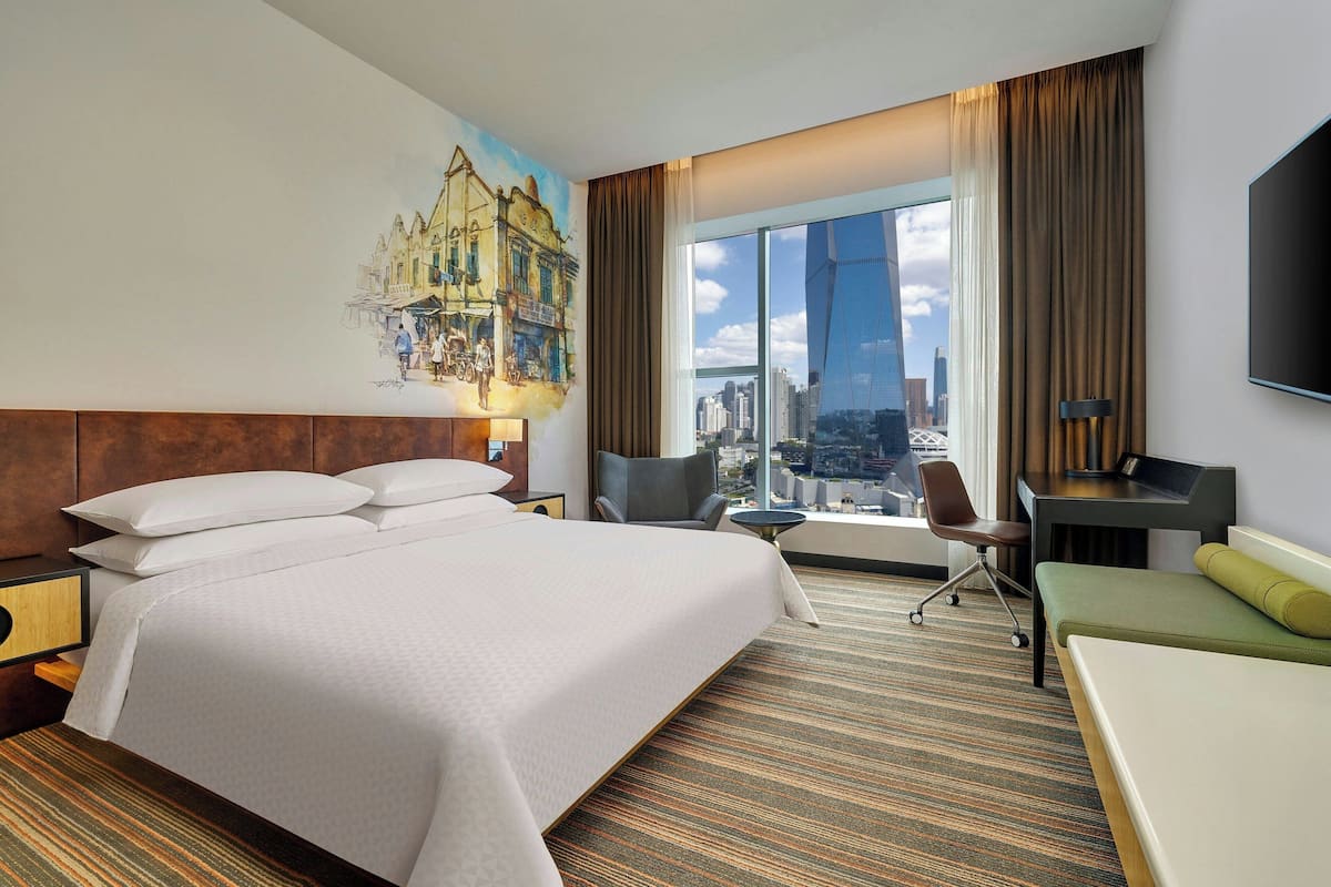 Habitación de hotel con una cama grande, un escritorio, una silla y vistas a un paisaje urbano a través de la ventana.