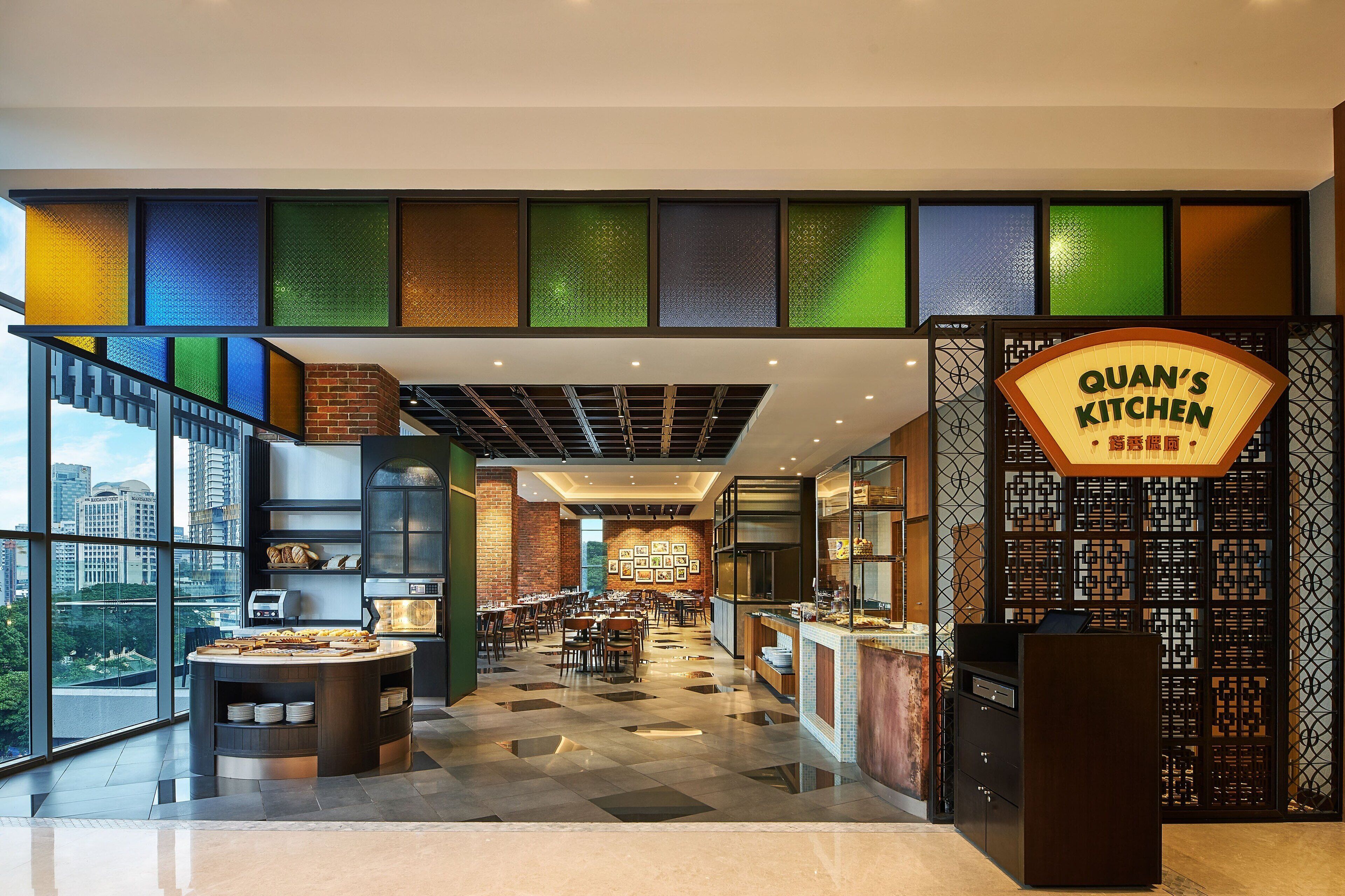 A modern restaurant with a central counter, multiple dining tables, and a view of the cityscape through large windows.