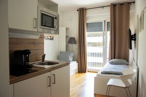 Una habitación compacta con kitchenette, una cama con almohada, una silla y un ventanal con cortinas.