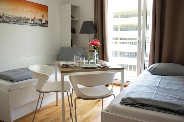 Une chambre d’hôtel avec un lit, une table à manger avec des chaises, une lampe, un vase de fleurs et un tableau représentant un paysage urbain accroché au mur.
