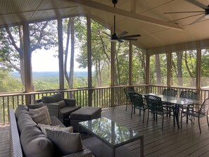 A covered outdoor seating area with a glass table and chairs, overlooking a forested landscape.