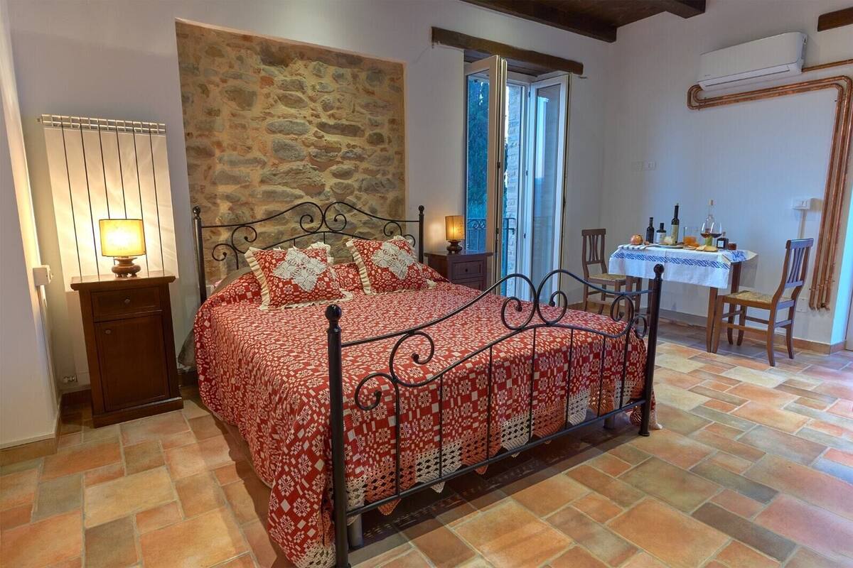 Une chambre avec un mur en pierre, un lit recouvert d’une couvre-lit à motifs, une table de chevet en bois, une table avec une nappe et une chaise.
