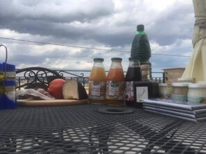 Una mesa con diversos alimentos y bebidas, incluyendo botellas de jugo y una botella de agua, en un balcón con vistas al cielo.