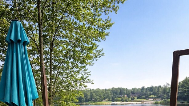 A wooden deck with a metal table and chairs, a blue umbrella, and a view of a lake with greenery in the background. - LAKEFRONT,Swim,enjoy&relax,7kayaks,SUP,paddlboat,firepit,PA,cabin,Fun,  (Tobyhanna)
