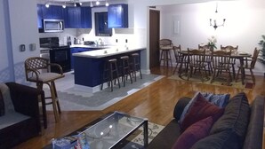 A modern kitchen with blue cabinets, a central island with stools, a dining area with wooden chairs, and a living room with a sofa and a glass coffee table. - LAKEFRONT,Swim,enjoy&relax,7kayaks,SUP,paddlboat,firepit,PA,cabin,Fun,  (Tobyhanna)