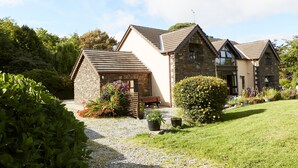 Une maison en pierre à deux étages, dotée d’un toit en pente, d’une allée gravillonnée et d’un jardin bien entretenu où poussent diverses plantes et fleurs.