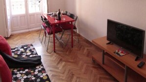 A living room with a wooden herringbone floor, a red dining table with chairs, a television on a wooden stand, and a wall-mounted shelf with decorative items. - Elegance Plaza Mayor with Balcony (Madrid)