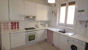 A compact kitchen with white cabinets, a gas stove, an oven, a washing machine, and a window with blinds. - Elegance Plaza Mayor with Balcony (Madrid)