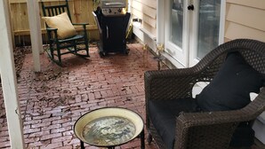 A covered patio area with a brick floor, a wicker chair, a small table, a grill, and a wooden fence.