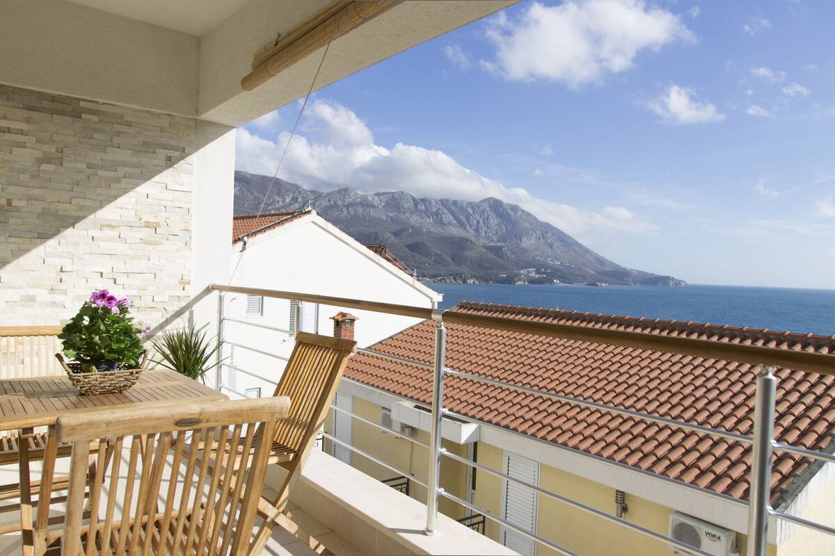 A balcony with a wooden table and chairs, a potted plant, and a view of the sea and mountains.