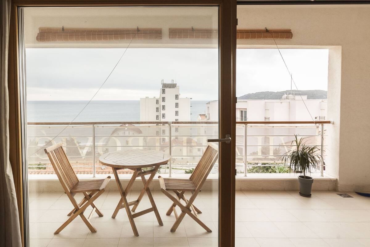 A balcony with a table and two chairs, a potted plant, and a view of the sea and buildings.