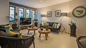 A modern living room with a sofa, two armchairs, a coffee table, and a TV. There are potted plants and framed pictures on the walls. - Icarus Suite Apartments (Rethymno)
