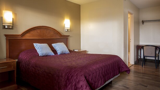 A bedroom with a large bed, two wall-mounted lamps, a nightstand, and a desk with a chair. - Viking Motel (Detroit)