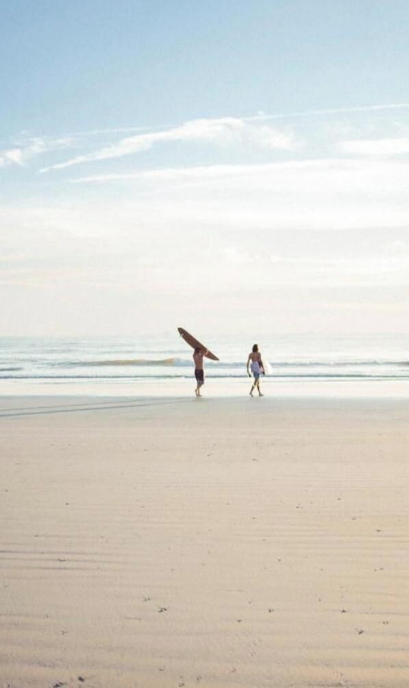 Una playa con una persona cargando una tabla de surf y otra caminando con una tabla.