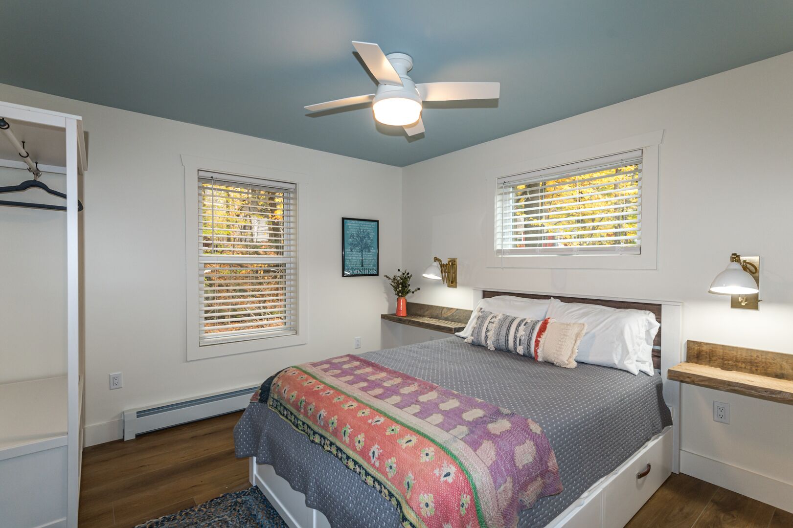 Une chambre à coucher comprenant un lit, un ventilateur de plafond, deux fenêtres équipées de stores, une table de chevet avec une lampe et une commode.