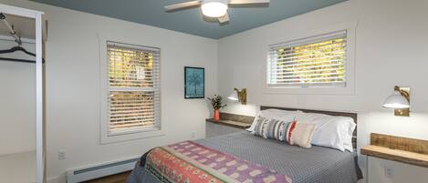 Une chambre à coucher comprenant un lit, un ventilateur de plafond, deux fenêtres équipées de stores, une table de chevet avec une lampe et une commode.
