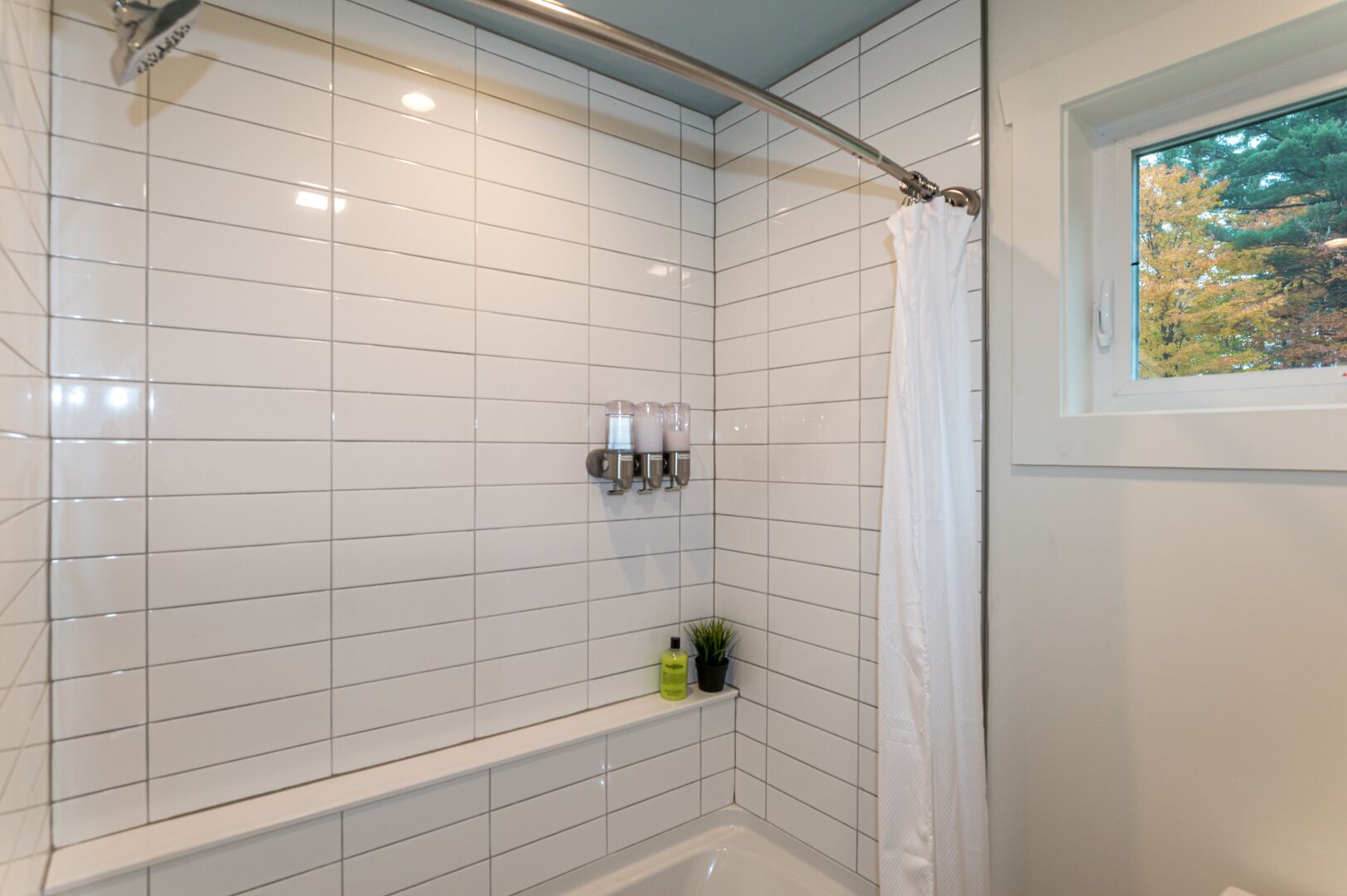 Ein Badezimmer mit weißen Fliesen, einer Duschvorhang, einem Fenster mit Blick auf Bäume und einem Ablagebereich für Toilettenartikel.