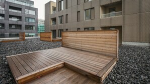 A modern outdoor seating area with a wooden bench and a small table, surrounded by a gravel surface, with a contemporary building in the background.