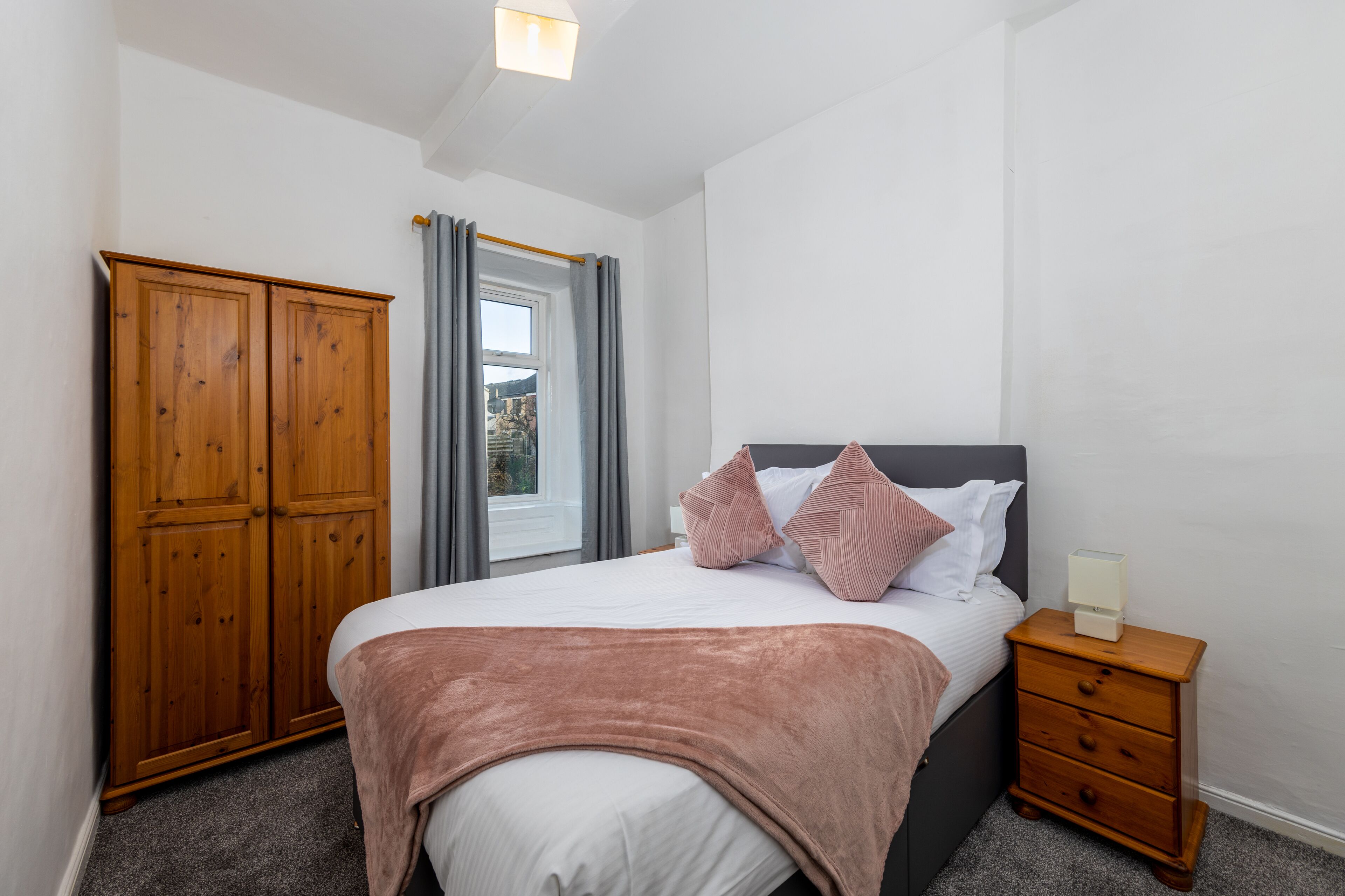 Een slaapkamer met een bed, een houten garderobe, een nachtkastje en een raam met gordijnen.