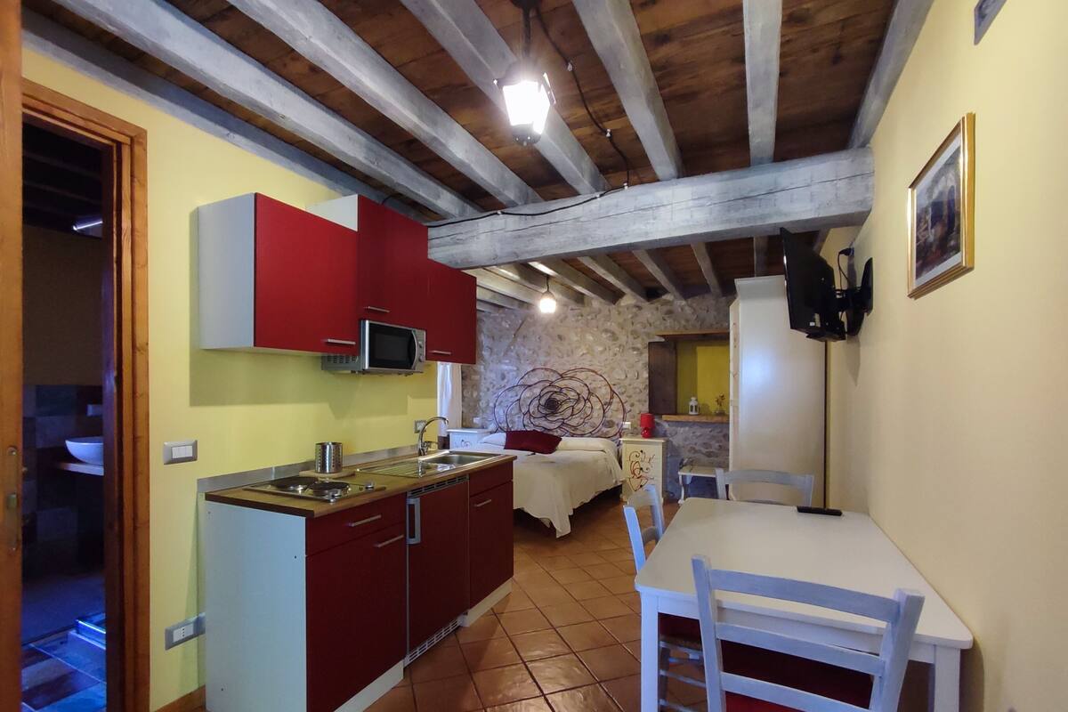 Une cuisine compacte avec des meubles rouges, un four à micro-ondes et un évier. On y trouve un coin repas avec une table et des chaises blanches. La pièce est dotée d’un mur en pierre et d’un plafond en bois.