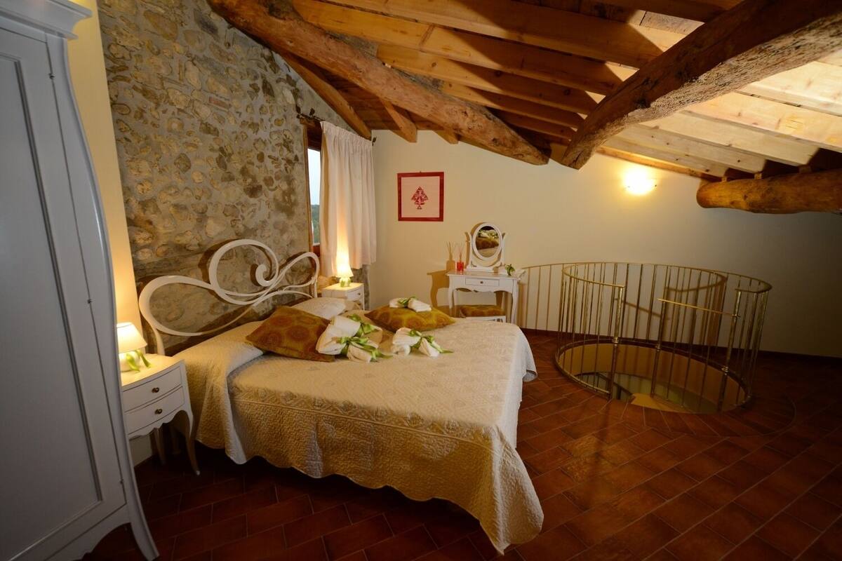 Une chambre avec un mur en pierre, des poutres en bois au plafond, un lit avec une tête de lit blanche, une table de chevet, une commode et une petite table ronde avec un miroir.