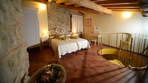 Ein Schlafzimmer mit Steinwand, einem Bett mit weißem Kopfteil, einem kleinen Nachttisch und einem Stuhl. Eine Holztreppe mit gelander ist ebenfalls zu sehen.