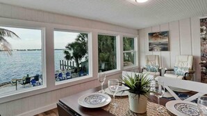 Ein Zimmer mit großen Fenstern mit Blick auf ein Gewässer, ein gedeckter Esstisch mit Tellern und Gläsern und eine Zimmerpflanze.