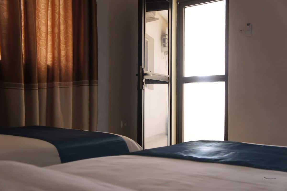 A hotel room with two beds, a large window with curtains, and a door leading to another room.