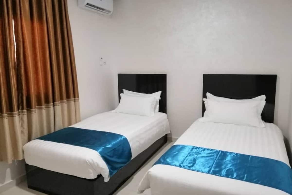 Two single beds with black headboards and white bedding, a brown curtain, and a white air conditioning unit on the wall.