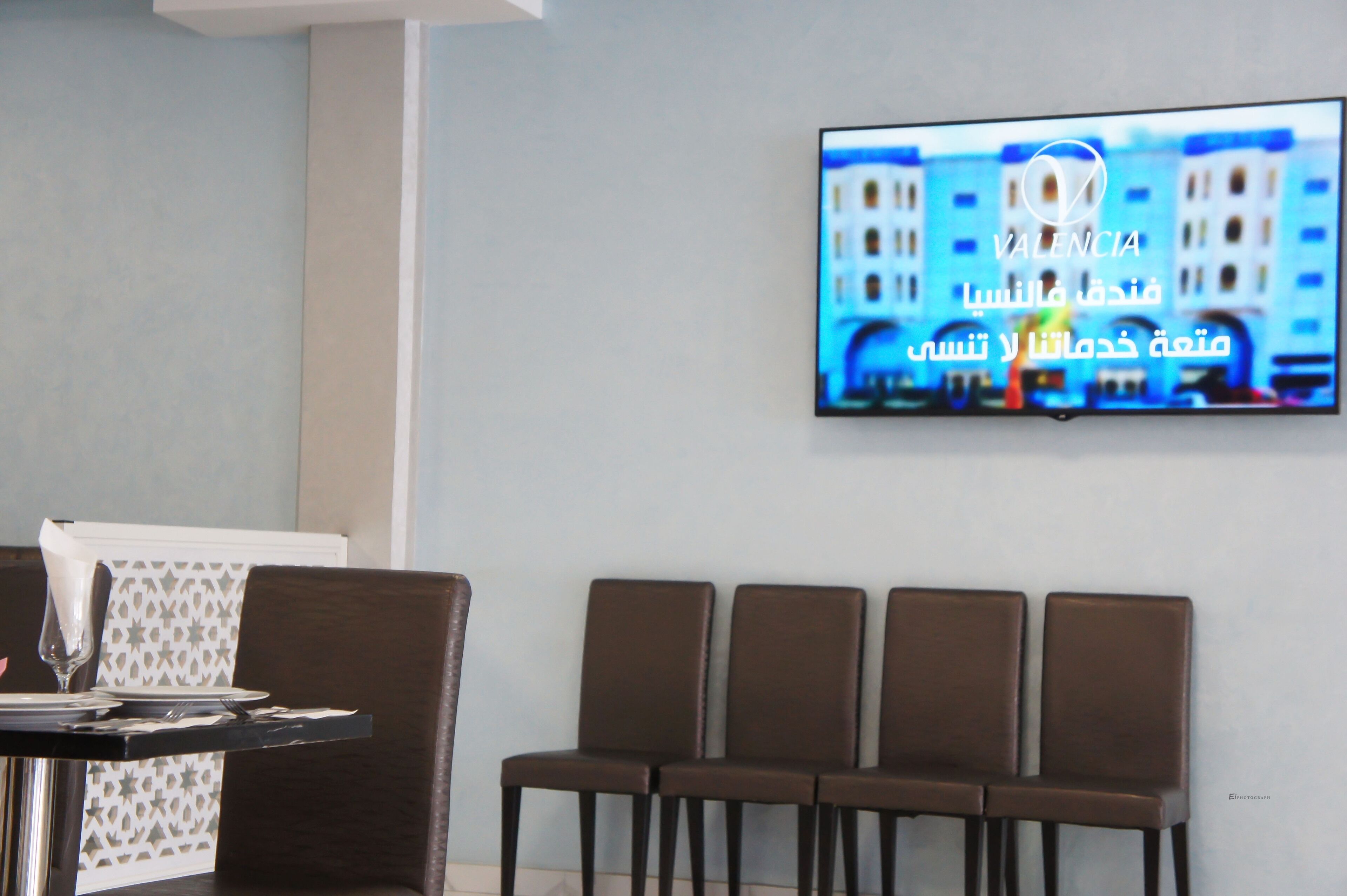 En hotellobby med en tv monterad på väggen som visar en stadsbild. Fyra bruna stolar är placerade i en rad.